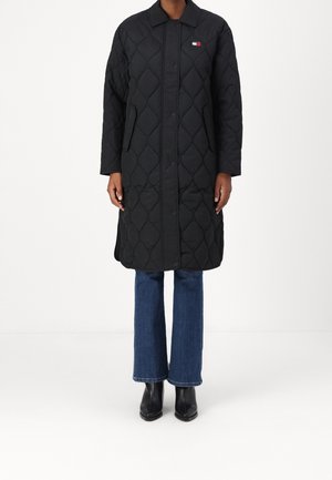 Persona con un abrigo largo acolchado negro con botones a presión, vaqueros azules y botines negros, de pie frente a un fondo blanco.