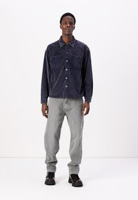 Cămașă din corduroy bleu marin cu butoni și două buzunare pe piept, purtată cu pantaloni din denim gri deschis și cizme negre cu șireturi. Design simplu, structurat.