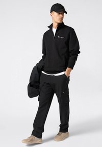 Pullover in pile nero con zip a un quarto, logo bianco, pantaloni cargo neri con tasche e sneakers beige. Il modello tiene un giubbotto nero.
