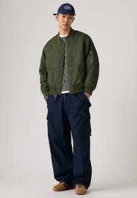 Φόρμα μπουφάν bomber σε ελιά με ριμπ μανσέτες και μέση, φορεμένο πάνω από γκρι πλεκτό πουλόβερ. Χαλαρό μπλε παντελόνι cargo και καφέ παπούτσια.