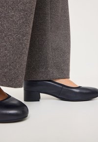 Scarpe in pelle nera a tacco basso con punta arrotondata e dettagli di cucitura, abbinate a pantaloni in tessuto grigio testurizzato.