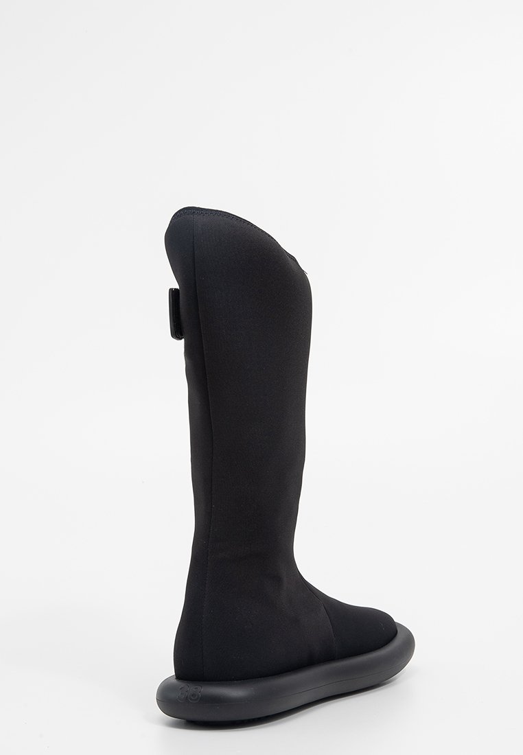 Czarne botki za kolano z gładkim, miękkim materiałem wierzchnim, zaokrąglonym czubkiem i teksturowaną gumową podeszwą. Posiadają tylną pętelkę do łatwego zakładania.