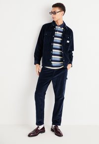 Giacca e pantaloni in velluto a coste blu navy con una maglietta a righe. Gli accessori includono occhiali da sole scuri e scarpe bordeaux. Calzini bianchi sono visibili.
