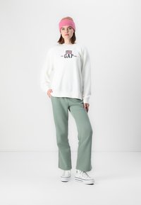 Biely polarový sveter s logom "GAP", v kombinácii s svetlozelenými nohavicami a bielymi teniskami. Model má na sebe ružovú pletenú čiapku.