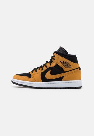 Kotníková teniska Nike Air Jordan v černé a hořčicově žluté semišové úpravě s černými tkaničkami a bílou stélkou na bílém pozadí.