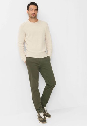 Man die een crèmekleurige trui, olijfgroene broek en camouflage sneakers draagt, staat met handen in de zakken tegen een witte achtergrond.