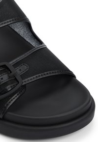 Svart sandal med en texturerad yta, bred rem med spänne och platt sula. Öppen tå-design med nätaccenter.