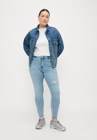 Denim jacket in dark blue, white t-shirt, light blue ripped jeans, and silver sneakers. Model stands with arms relaxed. Simple design.