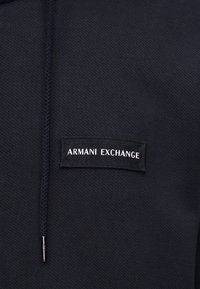 Κοντινό πλάνο σε ύφασμα ναυτικού μπλε με πλεκτό κορδόνι και ορθογώνια μαύρη ετικέτα με την επιγραφή "ARMANI EXCHANGE" σε λευκό κείμενο.