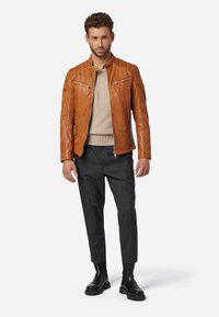 Bruine leren motorjack met geborduurde schouderdetails, gecombineerd met een beige trui en een op maat gemaakte antracietkleurige broek, en zwarte chelsea-boots.