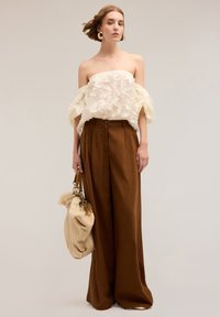 Top crema off-the-shoulder con ricami floreali testurizzati, abbinato a pantaloni marroni a gamba larga. Il modello tiene in mano una borsa beige con dettagli morbidi.