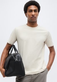 Άνδρας που φοράει απλό μπεζ μπλουζάκι κρατώντας μια μεγάλη μαύρη δερμάτινη τσάντα με λαβές, στέκεται μπροστά σε απλό φωτεινό φόντο.