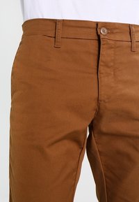 Bruine katoenen chino's met een gladde textuur, voorzien van een knoopsluiting, riemlussen en zijzakken, weergegeven op middeltorso tegen een lichte achtergrond.