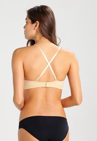 Beige podprsenka s křížovými ramínky, s jemnou texturou a zapínáním na háček. V kombinaci s černými bikinovými kalhotkami, s pohledem zezadu.