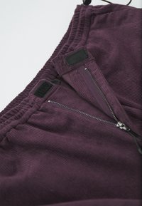 Pantaloni din corduroy mov, cu talie elastică, buzunare cu fermoar și accente velcro. Material texturat cu detalii verticale în formă de nervuri.