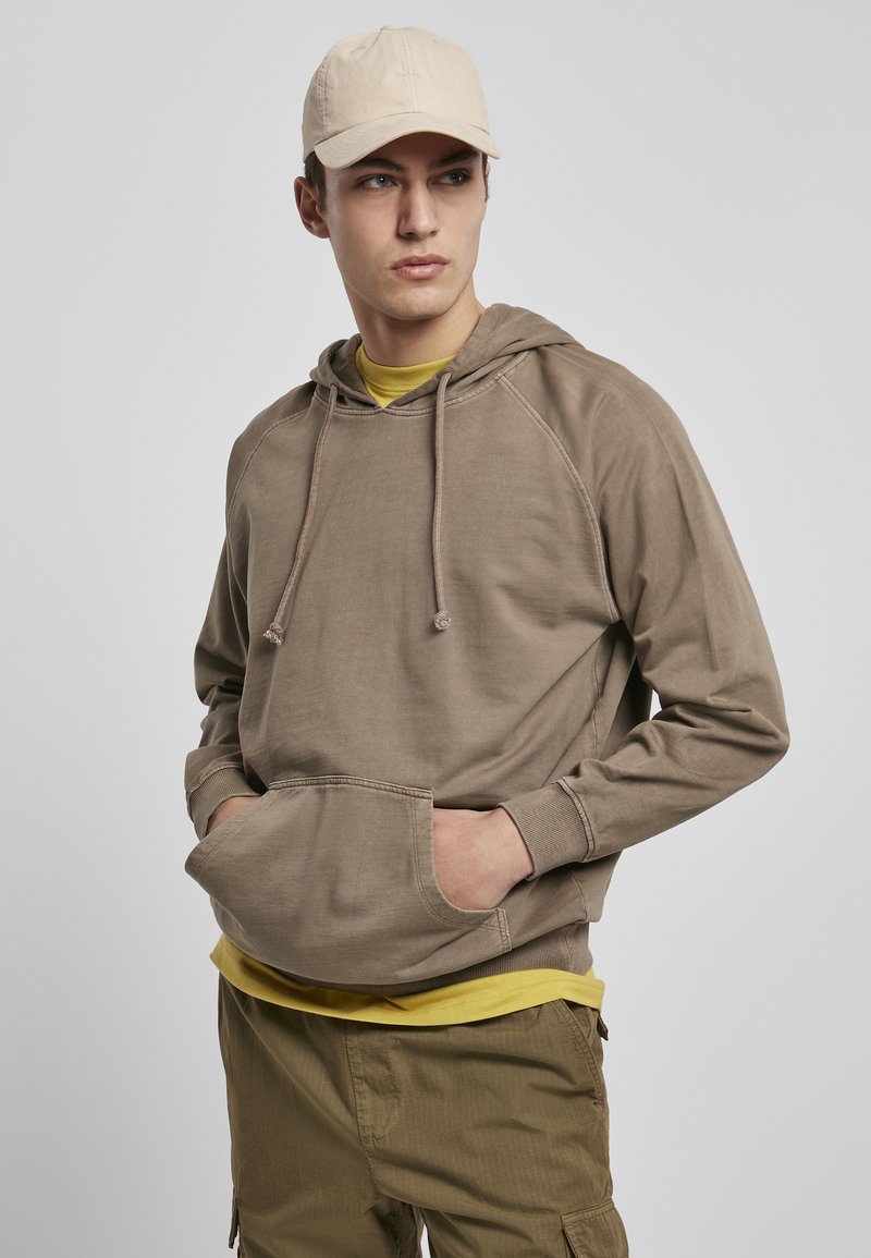 Felpa marrone con tasca a marsupio, cappuccio con cordino e polsini a coste, indossata su una maglietta gialla, abbinata a pantaloni beige.