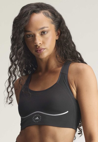 Kvinde med langt krøllet hår iført en sort sports-BH med en buet reflekterende stribe og et rundt logo på brystet.