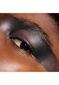 Nahaufnahme eines Auges mit aufgetragenem, dunklem, metallischem Lidschatten in einer auffälligen Form; die glatte Textur des Augenlids steht im Kontrast zu den Haaren in der Nähe der Augenbraue.