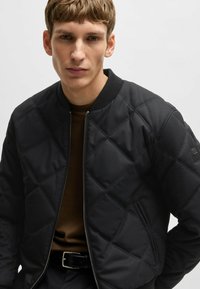 Quilted black bomber jacket with ribbed collar, zip closure, and side pockets, worn over a brown textured shirt.