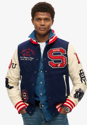 Navy and cream varsity jacket with red accents, featuring embroidered logos, snap buttons, and striped ribbed cuffs. Layered over a denim shirt.