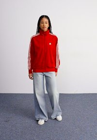 Giacca zip-up in velluto rosso con colletto alto e accenti bianchi a tre strisce sulle maniche, abbinata a jeans a gamba larga blu chiaro e sneakers bianche.