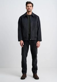 Ciemnoszara kurtka o strukturze konstrukcyjnej, z bocznymi kieszeniami i zamkiem błyskawicznym, noszona na ciemnozielonym swetrze i czarnych dżinsach. Brązowe botki za kostkę.