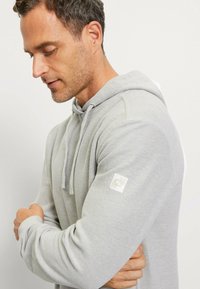 Grey hooded sweatshirt with textured fabric, long sleeves, and a small white logo patch on the left arm. Collected fit and layered design.