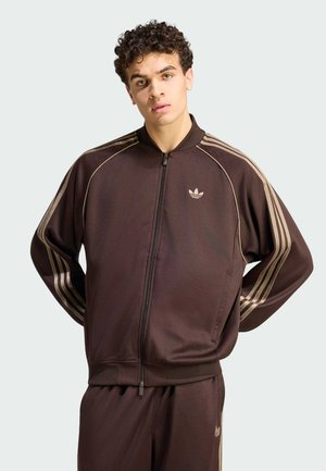 Młody mężczyzna z kręconymi włosami w brązowym dresie Adidas z beżowymi paskami, stoi z rękami za plecami na jednolitym tle.