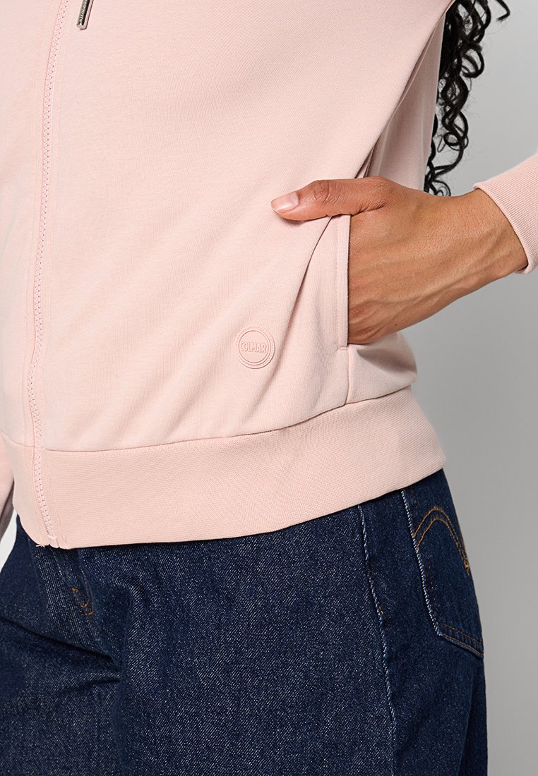 Giacca rosa chiaro con zip realizzata in tessuto morbido, dotata di tasche laterali e un logo circolare sull'orlo. Abbinata a jeans blu scuro.