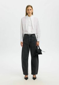 Naine seisab valge tausta taustal, seljas valge pika varrukaga pluus, millel on terav krae, mustad kõrge vöökohtadega teksad, mustad kontsad ning hoiab käes väikest musta käekotti.