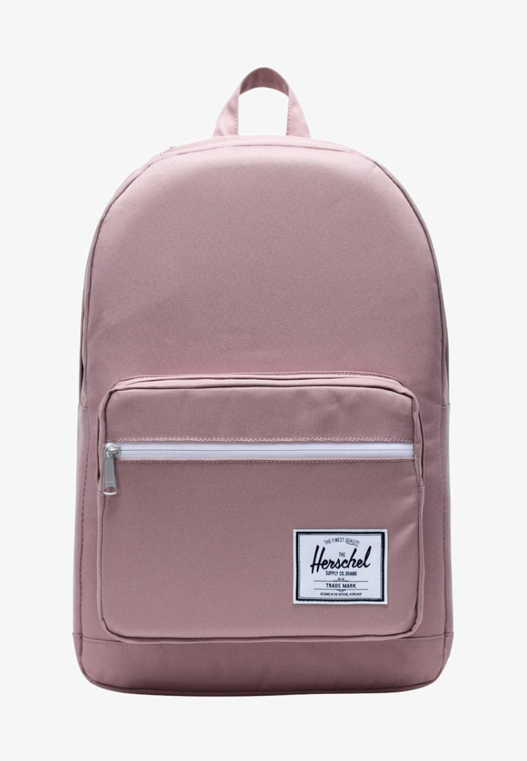 Sac à dos en tissu rose de forme arrondie, avec une poche zippée à l'avant, des accents blancs et une étiquette de marque. Il possède une poignée supérieure et des bretelles ajustables.