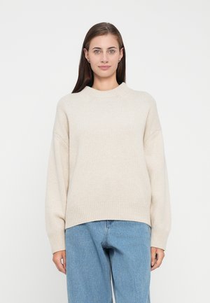 Beige stickad tröja med rund halsringning, ribbade ärmmuddar och en avslappnad passform, matchad med ljusblå jeans.