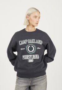 Donna bionda con capelli corti che indossa un felpa scura con la scritta "Camp Oakland Pennsylvania 1995", in piedi con le mani dietro la schiena.