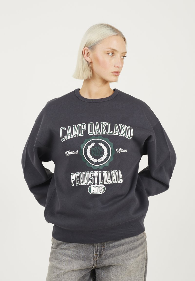 Donna bionda con capelli corti che indossa un felpa scura con la scritta "Camp Oakland Pennsylvania 1995", in piedi con le mani dietro la schiena.