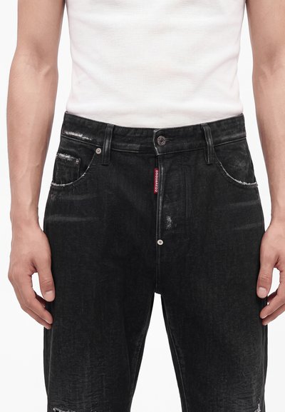 Homme portant un jean noir délavé et un t-shirt blanc sans manches, mains détendues sur les côtés, jean avec une petite étiquette rouge près de la fermeture éclair.