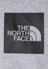 Tela gris con un parche negro cuadrado que lleva el texto blanco "THE NORTH FACE" y un logotipo estilizado con tres líneas curvas.