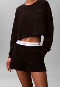 Czarna przycięta bluza z kapturem z przednią kieszenią, w połączeniu z czarnymi szortami z białym paskiem z logo. Gładka tekstura, casualowy design.