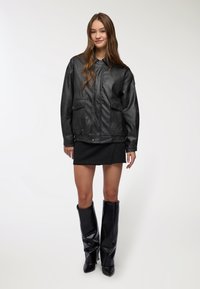 Giacca in ecopelle nera con colletto, due tasche frontali e vita elasticizzata. Indossata sopra a una minigonna nera e stivali alti fino al ginocchio.