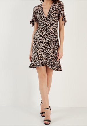 Kobieta w krótkiej sukience w panterkę z falbaniastymi rękawami i dołem, w połączeniu z czarnymi sandałami na obcasie z paskiem na kostce.