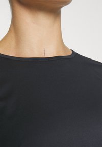 Svart tettsittende topp med rund hals, laget av glatt, elastisk materiale. En merkbar detalj er en liten linje på halsområdet.