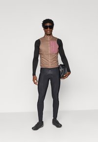 Kolesarska brezrokavnik v bež barvi z roza žepom, v kombinaciji s črnim dolgim rokavom in pajkicami. Model drži črno čelado in nosi temne sončne očala.