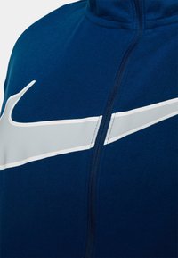 Giacca sportiva blu navy realizzata in tessuto liscio, con logo Nike bianco e chiusura frontale con zip. Il design con colletto alto migliora la copertura.