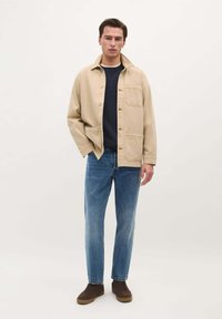 Beige jas met een knoopsluiting aan de voorkant, twee borstzakken, gedragen over een marineblauw shirt. Gecombineerd met blauwe rechte jeans en bruine schoenen.