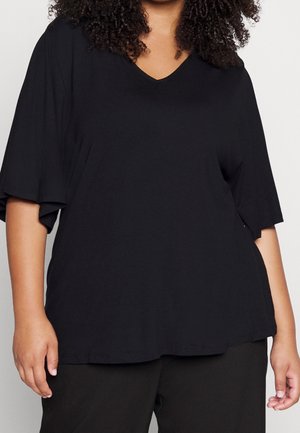 Personne aux cheveux bouclés portant un t-shirt noir ample à manches courtes et col en V, ainsi qu'un pantalon noir, sur un fond clair uni.