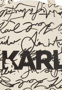 Bolsa de lona en crema, con texto garabateado en negro y un logo audaz de "KARL". Superficie texturizada con un patrón diverso de firmas.