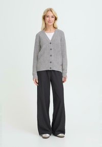 Cárdigan gris de punto con escote en V, siete botones en el frente y puños acanalados, acompañado de pantalones negros holgados y zapatos grises.