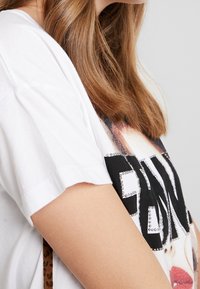 Camiseta blanca de algodón con mangas cortas, que presenta un estampado gráfico y letras negras decoradas con pequeñas tachuelas plateadas en la parte delantera.