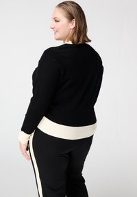 Zwarte trui met een crèmekleurige geribbelde tailleband en manchetten; heeft een aansluitend ontwerp en een zachte textuur; weergegeven van achteren.