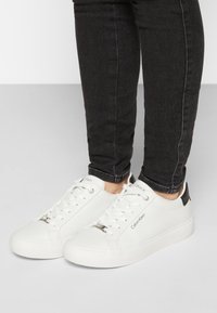 Baskets blanches à lacets avec une texture lisse, dotées d'un accent noir au talon et de détails métalliques sur la languette.