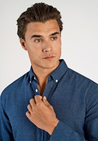 Camicia blu scuro con bottoni realizzata in tessuto strutturato, con collo allargato e dettagli con bottoni, parzialmente sbottonata al collo.